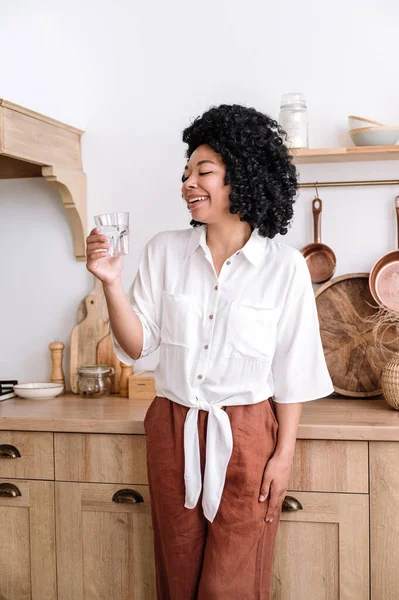 stock image Vertical shot of afro american woman in comfortable home clothes standing in kitchen and holding glass with water. Wooden furniture with accessories against female in casual wear