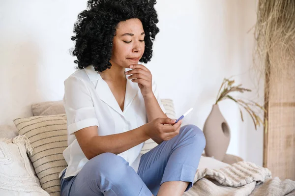 Future motherhood, fertility, infertility concepts. Disappointing afro american woman sitting on bed and holding pregnancy test. Positive or negative result. Female expecting baby.
