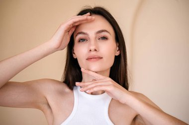 Mükemmel yüzüne dokunan genç bir kadının portresi. Açık tenli esmer, kameraya bakıyor. Kaş ve kirpik kaplama konsepti. Bej arkaplanda izole edilmiş model