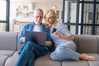İnternet sitesinden internet ve kredi kartı kullanan yetişkin bir çift. Karı koca evdeki kanepede oturuyor ve otelde rezervasyon yaptırıyor ya da tatil planlıyorlar.