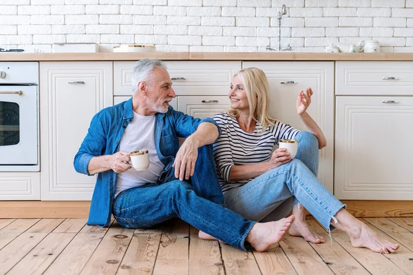 Mature husband and wife sitting on wooden floor and drinking morning coffee or tea together. Home comfort. Leisure weekend, dreaming, talking about plans.