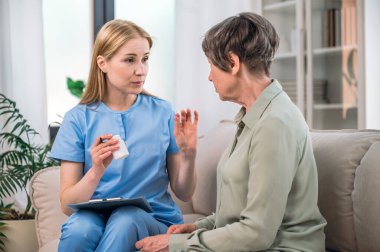 Mavi üniformalı kadın hemşire oturma odasında kanepede oturan yaşlı kadına antibiyotik hapı dozunu açıklıyor. Kronik hastalık, tıbbi tedavi, sağlık, yardım ve destek kavramı