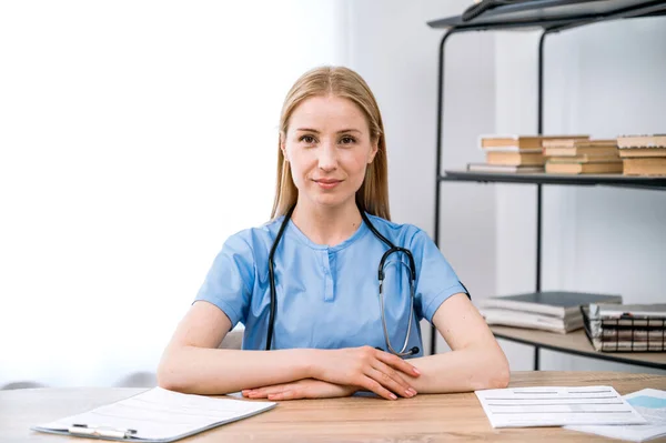 Portrait of professional general practitioner physician in blue uniform sitting behind table and waiting for patient in cabinet. Smiling female doctor looking at camera.