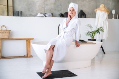 Beyaz bornozlu ve kafasında havlu olan genç bir kadın banyo küvetinin kenarında modern iç mimarisi ile oturuyor. Otel tuvaletinde bir kadın yüzüne dokunuyor ve duştan sonra dinleniyor. Vücut bakımı kavramı.