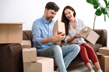 Mutlu genç çift kanepede oturuyor ve evdeki eşyaları kutulara dolduruyor. Yapışkan bant kullanarak. Kutuları yeni eve, daireye ya da daireye taşımak için hazırlayın..