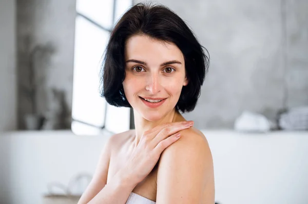 Young smiling woman after taking bath touching her shoulder, feeling soft smooth skin. Brunette female using moisturizer cream for body. Beauty and pampering procedure.