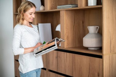 Modern İskandinav tarzıyla oturma odasındaki ahşap kitaplığın yanında duran, elinde dergi tutan, kitap okuyan, hafta sonlarını ev kütüphanesinde geçiren mutlu bir kadın. Hobi ve eğitim kavramı