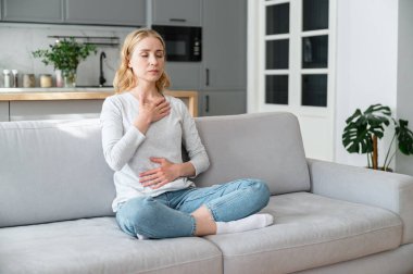 Göğüs ve karnında elleri olan, gözleri kapalı nefes alan, stresli bir durumda evde sakinleşen düşünceli genç bir kadın. Stresle başa çıkmak, iç dengeye ulaşmak.