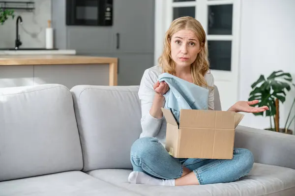 Confused woman receiving wrong parcel from internet store, checking size or color in cardboard box. Opening package at home. Frustrated female customer, bad client expirience, concepts