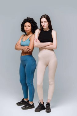 Gri arka planda yalnız duran sakin ve kendinden emin Afro-Amerikan ve Avrupalı kadınların dikey fotoğrafı. Sağlıklı yaşam tarzı, spor kıyafeti ve takım çalışması kavramı