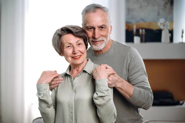 loving elderly man and his wife hugging and looking at camera in living room. happy mature couple give support and care each other. harmonious relationship concept
