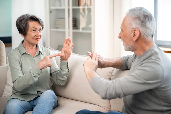 senior couple communicate in sign language while sitting on sofa at home. elderly deaf mute woman and man talking by hands gesture and smiling, understand each other