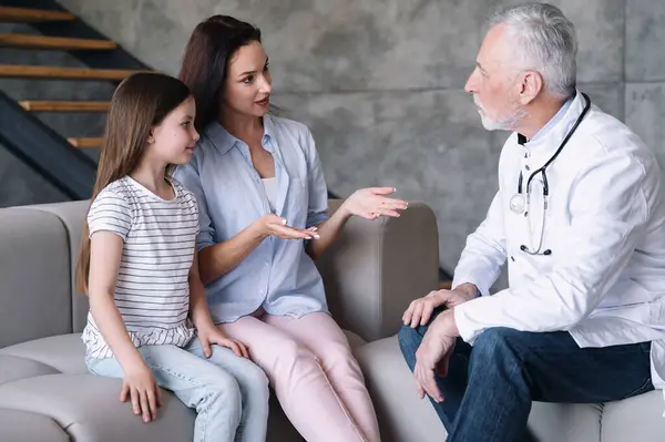Family doctor at home appointment. Woman sitting with daughter on couch, talking with physician and discussing about disease symptoms during consultation. Healthcare concept