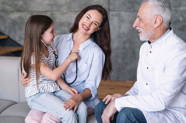 Домашний прием у педиатра. Дочь играет в доктора, проверяет мать, сидя на руках и слушать сердцебиение с помощью стетоскопа, мужчина и женщина смеются на диване в гостиной.