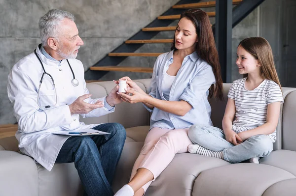 Family doctor in medical coat give pill to mother and daughter for disease treatment during home visit. Woman take medication jar in hand and listen pediatrician advice. Healthcare concept