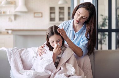 Bakıcı anne hasta kızının iyileşmesine yardım ediyor, evde kalıp destek oluyor. Kız kendini kötü hissediyor, burnu akıyor ve kanepede kareli bir şekilde otururken mendil kullanıyor.