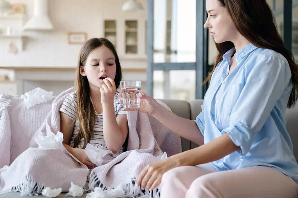 Caring woman bring her sick daughter medication pill and glass of water. Preteen girl feeling bad, have fever and runny nose, taking tablet while sitting wrapped in plaid on sofa. Healthcare concept