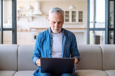 Odaklanmış orta yaşlı bir adam kanepede oturuyor, uzaktan kumandayla evde çalışıyor ve dizüstü bilgisayardan e-postaları okuyor. Erişkin erkek web sitelerini taramak ve internette bilgi aramak için bilgisayar kullanıyor
