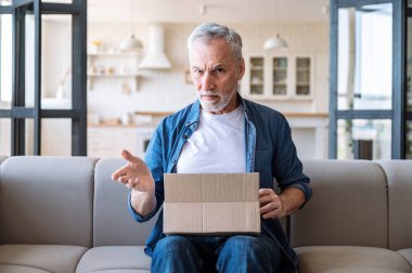 Memnun olmayan müşteri. Olgun bir adamın elinde yanlış ya da hasarlı siparişlerle dolu bir karton var. Kötü servis. Uzun teslimat, hata ya da hata yüzünden hayal kırıklığına uğramış. Evdeki kanepede oturan erkek..