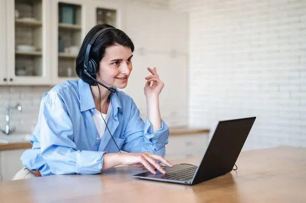 Young woman operator in headset working at call center online by laptop from home, sitting at desk and talking with client. Customer support service concept.