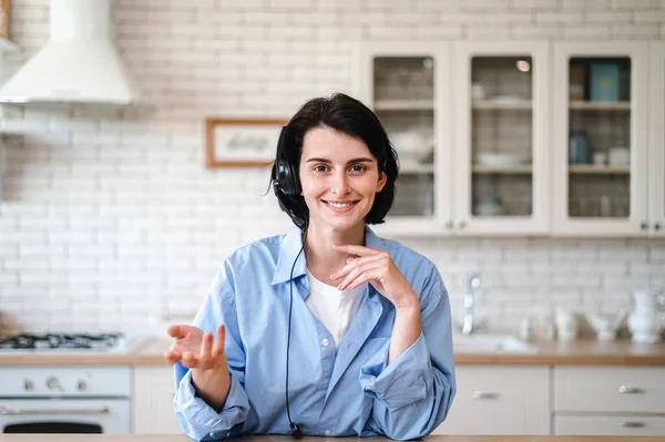 Smiling woman in headset talking with client by video call from home kitchen. Webcam view. Customer support service. Female have online interview with employer. Remote work concept.