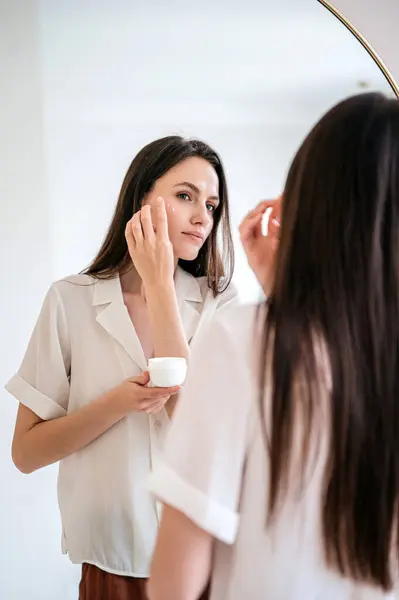 Woman holding cream jar in her hand, applying skincare product under eye. Beauty and skincare concepts. Portrait of young female model with fresh hydrated skin standing near mirror in bathroom