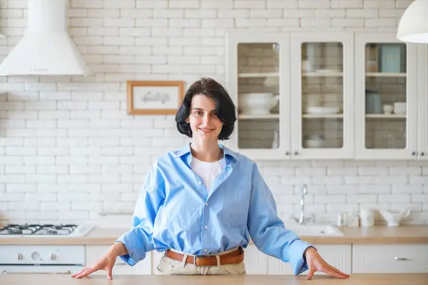 Smiling young woman in casual clothes standing near cooking table on kitchen background and looking at camera, make content for food blog. Female housewife start culinary masterclass