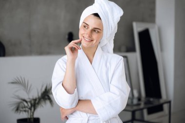 Beyaz bornoz ve havlu giymiş genç bir kadın duştan sonra banyoda ayakta duruyor ve sabah prosedürleri ve gülümsüyor, ferahlıyor. Ev kaplıcası ve güzellik ayini konsepti