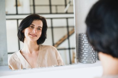 Genç, güzel bir kadın aynada yansımaya bakıyor, gülümsüyor, temizlendikten sonra sağlıklı cildinin tadını çıkarıyor ve banyoda dururken güzellik prosedürlerini uyguluyor. Kişisel bakım kavramı