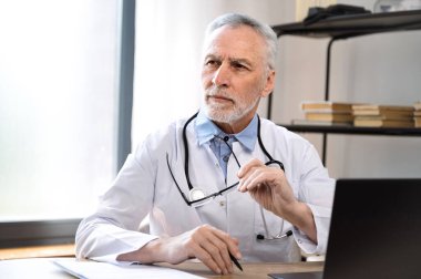 Beyaz üniformalı, düşünceli, olgun bir erkek doktor hastane dolabında oturmuş uzaktan bakıyor ve teşhisi düşünüyor. Düşünceli hekim klinikte karar verdi