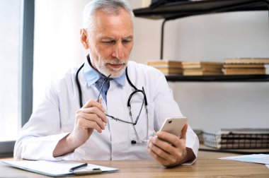 Beyaz önlüklü olgun bir doktor hastaya mesaj atıyor, online danışmanlık yapıyor. Uzaktan çalışma için sağlık uygulaması kullanan pratisyen, tıbbi kartları kontrol eden.