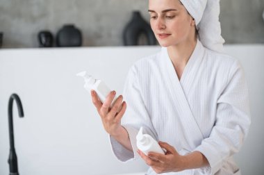 Bornozlu bir kadının cilt bakımı ve cilt bakım ürünlerini seçmesi. Pompalı şişeler. Günlük rutinler için organik, doğal kozmetik ürünler. Güzellik ve sağlık konsepti.