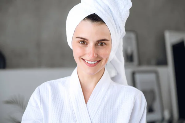 Portrait of happy woman with healthy skin wearing white bathrobe and towel on head, looking at camera and smile while standing at bathroom. Young female after morning beauty ritual. Home spa concept