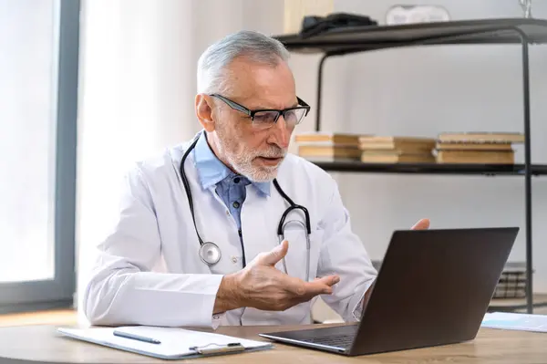 Concentrated professional male doctor in glasses and uniform looking at laptop screen, making video call and giving online virtual consultation to patient from hospital office
