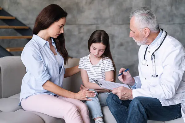 Girl patient upset about diagnosis, sitting on sofa with head down, mother supporting daughter and doctor explain treatment plan. Pediatrician at home appointment. Healthcare concept