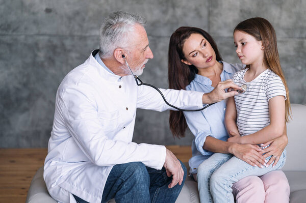 Врач-педиатр в медицинском пальто осматривает больного ребенка, проверяя легкие стетоскопом во время домашнего приема. Расстроенная девушка сидит в материнской руке, чувствует себя больным и имеет симптомы респираторной инфекции