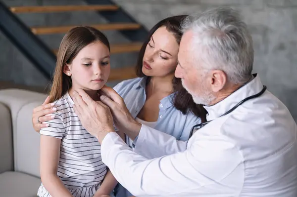 Friendly caring pediatrician examining kid, touching lymph nodes. Doctor checking sad girl for respiratory infection, virus, flu at home while her mother support little patient daughter