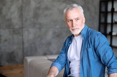 Beyaz tişört ve kot gömlek giyen, kendinden emin orta yaşlı bir adamın portresi. Oturma odasında kanepede otururken başka yere bakıyor ve projeyi düşünüyor. Akıllı serbest çalışan erkek evde çalışıyor.