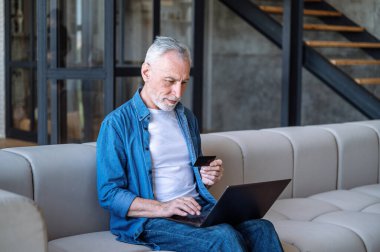 Laptop kullanan olgun bir adam, internet bankacılığına giriş yapan, klavyede şifre yazan, banka kartındaki bakiyeyi kontrol eden, evdeki koltukta oturan. İnternetten alışveriş.