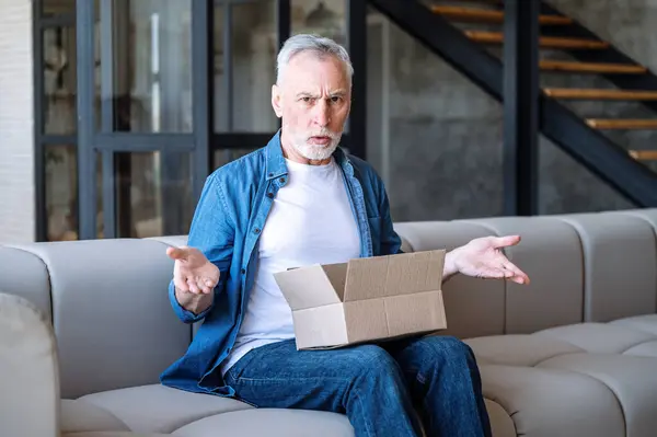 Annoyed senior man unpacking delivered cardboard parcel with order. Negatively surprised, confused, angry client received wrong or damaged item. Unhappy male customer with postal delivery