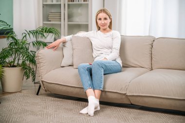 Rahat bir kanepede oturan rahat bir oturma odasında dinlenen rahat elbiseli genç bir kadın. Modern apartmandaki koltukta sakin bir kadın kameraya bakıyor ve gülümsüyor..