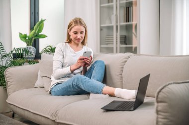 Mutlu kadın rahat bir kanepede dinleniyor ve evdeki internetli modern aletleri kullanıyor. İş görüşmesi için akıllı telefondan mesaj yazan serbest yazar, dizüstü bilgisayarın yanında oturur ve çevrimiçi video izlerdi.