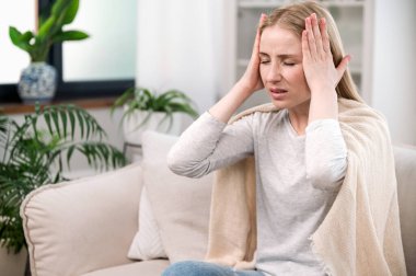 Akut baş ağrısı çeken stresli genç bir kadın ekoseye sarılı bir şekilde kanepede otururken elleriyle tapınakları tutuyor. Mutsuz kadın migrenden muzdarip, sağlık probleminden yorgun..