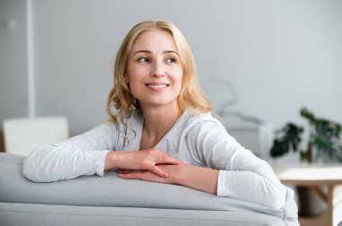 Sağlıklı gülümseyen, neşeli, mutlu, genç bir kadın portresi rahat bir oturma odasında rahat bir kanepeye oturup yan yan bakıyor ve hafta sonları evdeki sabahının tadını çıkarıyor. Eğlence, rahatlama kavramları