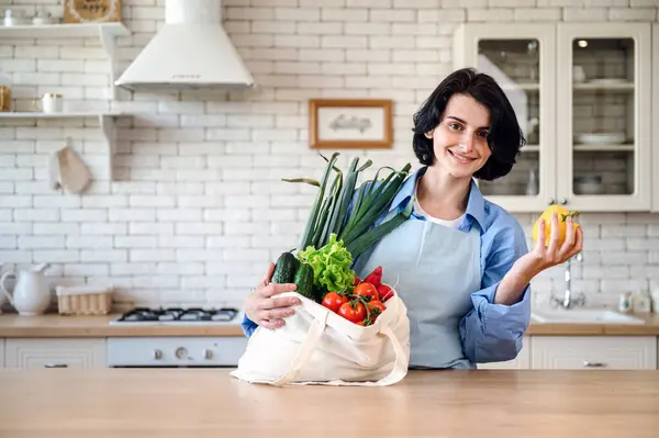 Happy woman at home getting groceries out of shopping bag. Ordering from internet fresh organic vegetables and greens. Kitchen interior. Food delivery concept