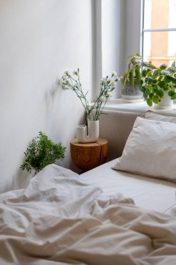 Parlak zarif iç mekanın ayrıntıları rahat yatak odasında. Ahşap sehpa, seramik vazo ve çiçeklerle rahat yatağın yanında keten yatak. Pencere kenarında saksı bitkileri