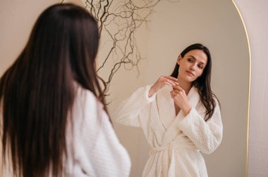 Beyaz bornozlu kadın duştan sonra banyoda dikilip aynaya bakıyor ve saç hasarını kontrol ediyor. Saç bakımı, güzellik ve sabah rutini kavramları