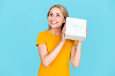 Gülümseyen genç bir kadın, mavi arka planda tek başına dururken hediyesini alıp, ellerinin arasında tutarak hayal kuruyor. Paketi olan neşeli, düşünceli bir kadın. Tatil kavramı.