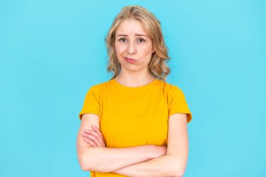 Studio shot of woman with crossed arms, looking at camera, having doubtful and indecisive expression. Confused young female posing isolated on blue background. clipart
