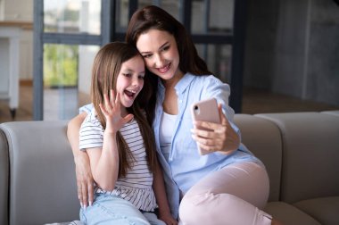 Kanepede oturan, annesini kucaklayan, akıllı telefona bakan, el sallayan ve video görüşmesiyle konuşan gülümseyen kız. Neşeli kadın evde kızıyla vakit geçiriyor. Mutlu aile kavramı